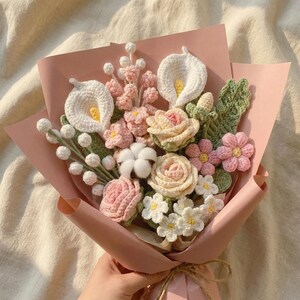 Peut inclure: Un bouquet artisanal de fleurs au crochet dans les tons de blanc, rose et cr&egrave;me, envelopp&eacute; dans du papier rose. Le bouquet comprend des arums, des roses et d'autres &eacute;l&eacute;ments floraux, attach&eacute;s avec de la ficelle.