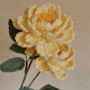 May include: A handmade crochet flower featuring two large, pale yellow blooms with a bright yellow centre. The flower has green leaves and stems. The petals are detailed, and the overall effect is soft and textured.