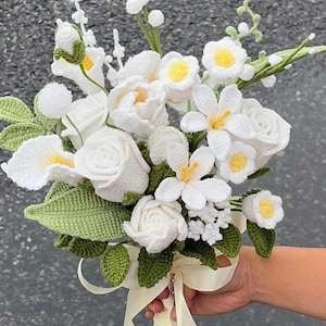 Op de afbeelding: Een handgemaakt gehaakt boeket met witte bloemen met gele centra en groene bladeren. Het boeket bevat rozen, calla's en andere bloemelementen, vastgebonden met een crèmekleurig lint. De bloemen zijn ingewikkeld vervaardigd met gedetailleerde steken.