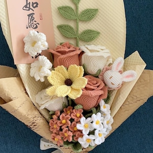 以下が含まれることがあります： ピンク、白、黄色の糸で編まれた花束と、小さな編みぐるみウサギ。花束は茶色の紙で包まれ、金色の文字で「愛はより良い」と書かれた白いリボンで結ばれています。