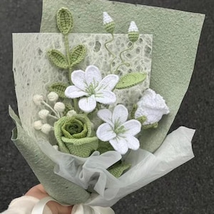 Puede incluir: Un ramo artesanal de flores de ganchillo, con flores blancas, hojas verdes y una rosa verde. El ramo está envuelto en papel verde claro y atado con una cinta blanca. Un arreglo floral único y hecho a mano.