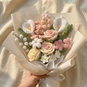 Peut inclure: Un bouquet de fleurs crochet&eacute; &agrave; la main avec des callas blanches, des roses roses et d'autres fleurs dans des tons de rose, de jaune et de blanc. Le bouquet est envelopp&eacute; dans du papier cr&egrave;me et nou&eacute; avec un ruban blanc.