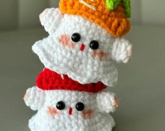May include: A stack of two white crochet ghost figures, one wearing a red scarf and the other a pumpkin hat. The ghosts have black bead eyes and pink blush. The pumpkin hat is orange with a green stem.