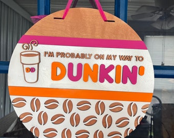 Può includere: Un cartello rotondo in legno con un design a strisce rosa, arancio e bianco. Il cartello dice "I'm probably on my way to Dunkin'" con una grafica di una tazza di caffè. Lo sfondo è un motivo bianco di chicchi di caffè.