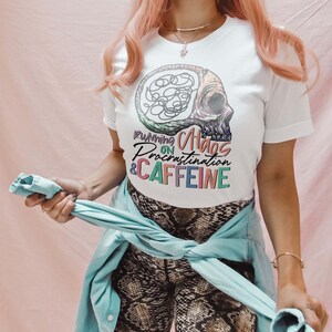 K&ouml;nnte beinhalten: Wei&szlig;es T-Shirt mit einer bunten Grafik eines Sch&auml;dels mit einem Gehirn darin. Der Text "Running on Chaos, Procrastination & Caffeine" ist auf das Shirt gedruckt.