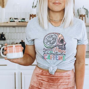 K&ouml;nnte beinhalten: Ein graues T-Shirt mit einer bunten Grafik eines Sch&auml;dels mit einem Gehirn darin. Der Text auf dem Shirt lautet "Running on Chaos, Procrastination & Caffeine".