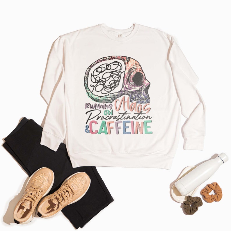 K&ouml;nnte beinhalten: Wei&szlig;er Pullover mit einem bunten Totenkopf-Motiv und dem Text "Running on Chaos, Procrastination & Caffeine".