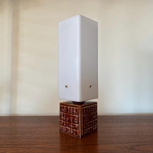 Puede incluir: Una lámpara de cerámica blanca con una pantalla cuadrada y una base de cerámica marrón con un patrón geométrico.