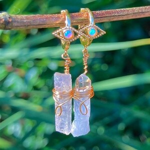 May include: Gold-colored hoop earrings with an eye design and dangling clear crystal quartz. The crystals are wrapped in gold wire. The earrings are hanging from a brown twig against a blurred green background.