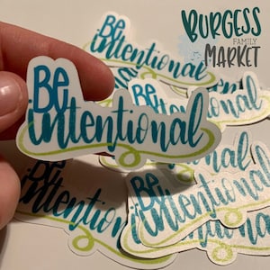 May include: A collection of stickers with the phrase "Be Intentional" in teal script. The stickers have a white background with a light green border. The Burgess Family Market logo is in the upper right corner.