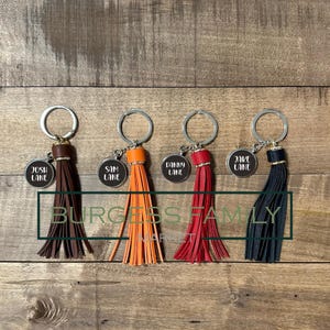 May include: Four keyrings with leather tassels in brown, orange, red, and black. Each keyring has a tag with a name: Josh Lane, Sam Lane, Danny Lane, and Jake Lane. The text "BURGESS FAMILY" is also visible. The keyrings have silver rings.