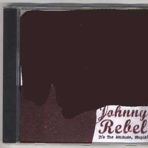 Puede incluir: Una caja de CD con una cubierta marrón oscuro, parcialmente oscurecida por una forma grande e irregular. La esquina inferior derecha revela el texto "Johnny Rebel" en una fuente estilizada, con "It's The Attitude, Stupid!" debajo.