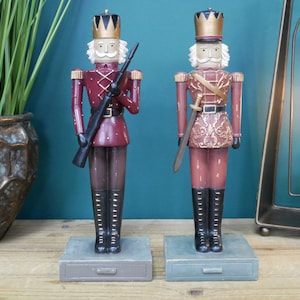May include: Two red and gold painted wooden nutcracker figurines standing on gray bases with a drawer. The nutcracker on the left is holding a rifle and the nutcracker on the right is holding a sword.