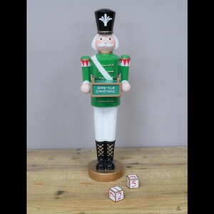 Può includere: Un schiaccianoci di Natale verde e bianco con un cartello che dice "Days Til Christmas". Lo schiaccianoci tiene il cartello nelle sue mani. Ci sono tre blocchi di legno con numeri su di essi, 1, 2 e 5.