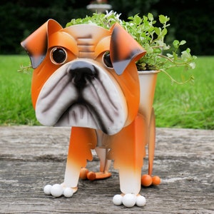 Puede incluir: Macetero de bulldog naranja y blanco con una planta verde en su interior. El macetero está hecho de metal y tiene un diseño realista.