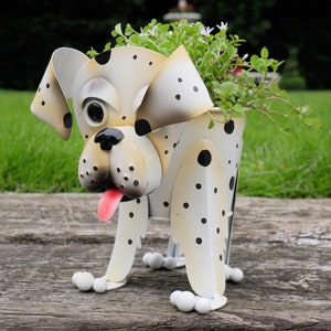 Puede incluir: Macetero de metal blanco con forma de dálmata con manchas negras. El macetero tiene una cabeza grande y redonda con ojos grandes y una lengua rosa que sobresale. El macetero está de pie sobre cuatro patas y tiene una maceta pequeña para plantas en la espalda.