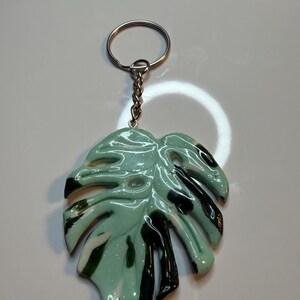 Peut inclure: Un porte-clés avec une breloque brillante en forme de feuille de monstera, fabriquée en résine marbrée vert menthe, blanc et vert foncé avec des paillettes subtiles. La breloque est fixée à un anneau en métal argenté par une courte chaîne assortie.