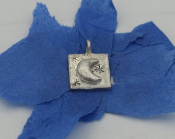 Puede incluir: Un dije de plata con un diseño de luna creciente y estrellas. El dije es de forma cuadrada y tiene una superficie texturizada.