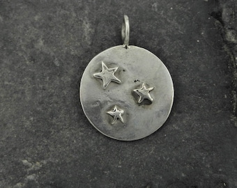 May include: A silver charm with three small star shapes embossed on a round disc. The charm has a loop at the top for attaching to a necklace or bracelet.
