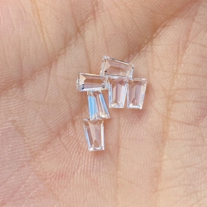 Peut inclure: Un groupe de six pierres précieuses rectangulaires transparentes et facettées. Les pierres précieuses sont disposées en un tas lâche sur une surface brun clair.