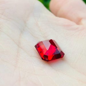 May include: A faceted, rectangular-cut gemstone with a deep, vibrant red color. The stone features multiple polished surfaces that reflect light, creating a brilliant appearance with sharp edges and a clear, transparent quality throughout the gem.