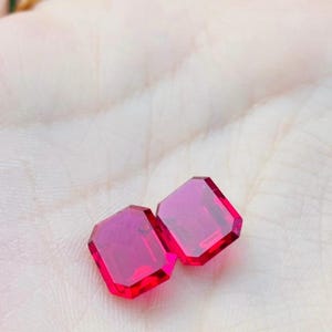 May include: Two vibrant, deep red gemstones cut in a rectangular emerald shape with faceted surfaces that reflect light. These loose, polished stones feature sharp, clean edges and a brilliant, saturated crimson color throughout their transparent structure.