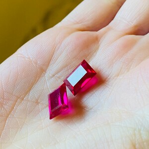 May include: Two red rectangular gemstones, one slightly larger than the other, are displayed on a hand.