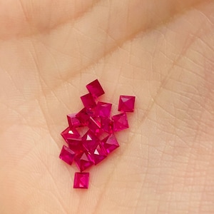 May include: A collection of small, square-shaped, ruby red gemstones. The gemstones are scattered on a person's hand.