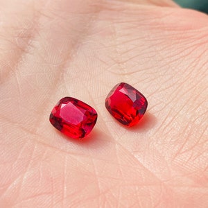 Könnte beinhalten: Zwei rote Edelsteine, geschnitten in einer quadratischen Form mit abgerundeten Ecken, werden auf einer Hand gezeigt. Die Edelsteine sind von einer tief roten Farbe und haben eine glänzende Oberfläche.