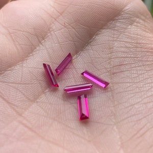May include: Five small, rectangular, pink gemstones on a hand.