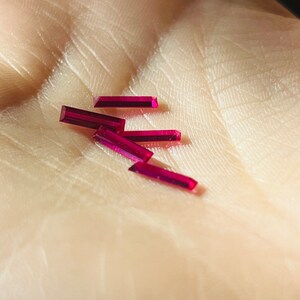 May include: Five small, rectangular, ruby red gemstones on a person's hand.