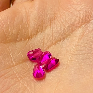 Puede incluir: Cinco gemas facetadas de color rosa intenso de varias formas descansan en la palma de una mano. Las gemas son de un color magenta vibrante y reflejan la luz. La mano es de piel clara y tiene anillos de oro en los dedos.