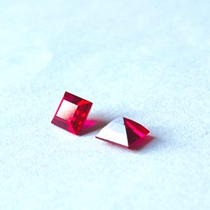 Könnte beinhalten: Zwei facettierte, quadratisch geschliffene, rote Edelsteine. Einer ist ein perfektes Quadrat, der andere ein Dreieck mit flacher Oberseite. Die Edelsteine sind hochreflektierend, mit einer hellen, satten Farbe. Der Hintergrund ist eine helle, strukturierte Oberfläche.