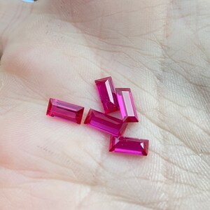 Pode incluir: Seis gemas retangulares cor-de-rosa, provavelmente rubis, são exibidas em uma mão. As gemas são facetadas e têm um acabamento brilhante.