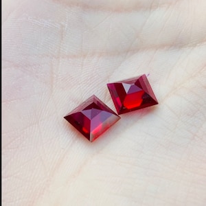 May include: Two red square-shaped gemstones on a white surface.