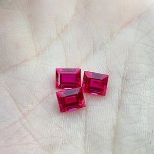 Può includere: Tre pietre preziose rettangolari, sfaccettate, di colore rosso rubino su una superficie bianca.