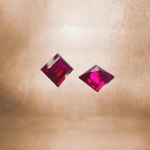 Puede incluir: Dos piedras preciosas de forma cuadrada, de color rojo intenso. Las piedras preciosas están facetadas y tienen un acabado brillante y pulido.