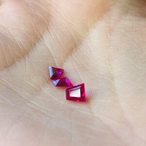 May include: Three red triangular gemstones on a hand. The gemstones are faceted and have a shiny finish.