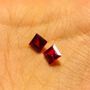 Peut inclure: Deux pierres précieuses carrées de couleur rouge foncé sur une surface brun clair.
