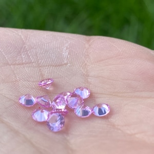 Könnte beinhalten: Eine Sammlung kleiner, rosafarbener, facettierter Edelsteine, wahrscheinlich Zirkonia, die auf einer Hand verstreut sind.