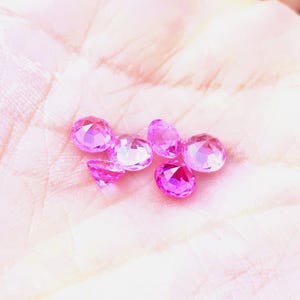May include: A group of six pink gemstones, likely sapphires, are displayed on a hand. The gemstones are oval-shaped and have a faceted cut.