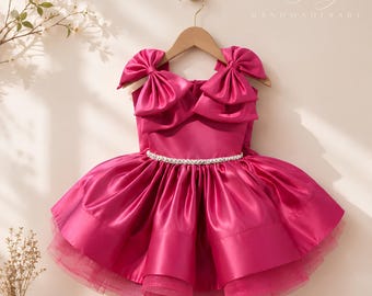 May include: A vibrant fuchsia dress hanging on a wooden hanger, featuring large bows on each shoulder, a fitted bodice, and a full, pleated skirt with a beaded waistband. The text Jolly HANDOMADIBABY is visible in the upper right corner.