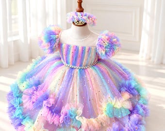 May include: A multi-colored tulle dress featuring a square neckline, puffed sleeves, and a full, layered skirt with ruffled edges. The fabric displays a rainbow gradient pattern with gold star accents and a waistline embellished with silver beads.