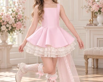Puede incluir: Un vestido rosa claro sin mangas con escote cuadrado y falda acampanada que presenta volantes de encaje blanco superpuestos en el dobladillo. El conjunto incluye calcetines altos de encaje blanco con lazos rosas y zapatos claros con hebilla.