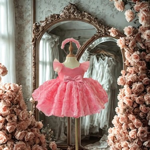 May include: A pink tulle dress with a rose pattern, bow, and matching headband displayed on a mannequin. The dress has a layered skirt and flutter sleeves. The background includes a mirror and floral arrangements.