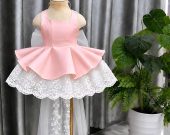 May include: A sleeveless dress featuring a solid light pink bodice with a flared peplum waist, layered over a white lace skirt with a scalloped hem and a sheer white tulle train extending from the back, displayed on a mannequin.