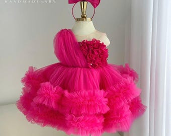 May include: A bright fuchsia tulle dress featuring a one-shoulder design, a large cluster of fabric petals on the bodice, and a voluminous multi-layered ruffled skirt. A matching fuchsia bow headband is displayed above the dress. Text reads Jelly HANDMADEBABY.