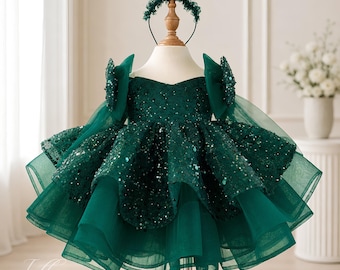 Op de afbeelding: Een donker smaragdgroene jurk met een lijfje met lovertjes en een meerlaagse tule rok met volumineuze ruches. De jurk heeft off-shoulder mouwen met grote strikaccenten en wordt getoond met een bijpassende donkergroene hoofdband met kralen.