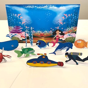 Puede incluir: Un conjunto de recortes de papel de colores de criaturas marinas, que incluyen una ballena, peces, una sirena, un cangrejo, un delfín, un pez globo y un submarino. Los recortes están dispuestos sobre una superficie blanca frente a un fondo de océano azul y verde.