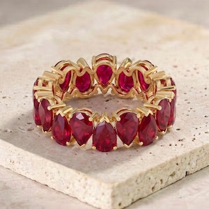 Könnte beinhalten: Ein goldener Eternity-Ring mit einem durchgehenden Band aus leuchtend roten, birnenförmigen Edelsteinen. Die Steine sind in einer Krappenfassung gefasst und bilden eine kreisförmige Anordnung, die die tiefrote Farbe und die Facetten betont.